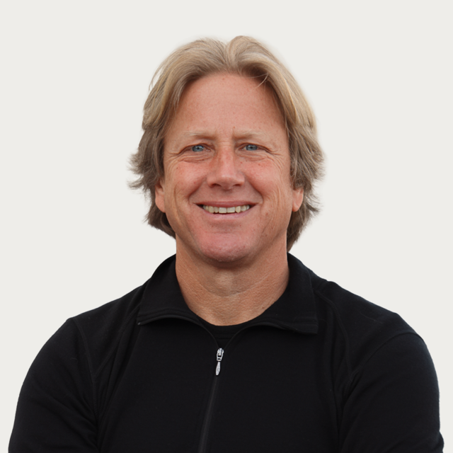 A middle-aged man with blonde hair wearing a black zip-up jacket, smiling at the camera against a plain white background.