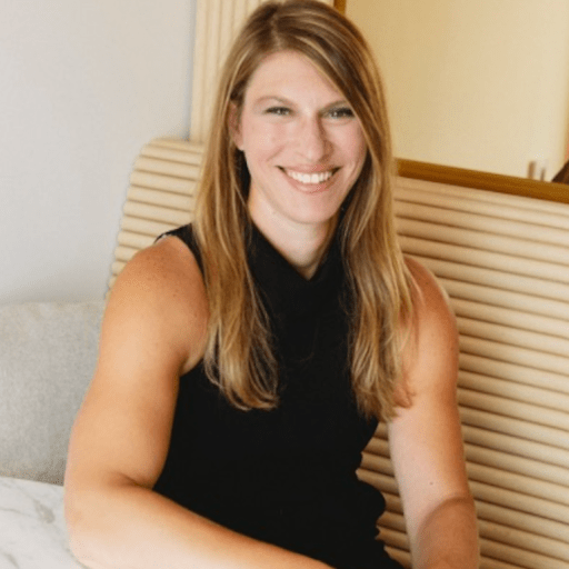 A woman with long blonde hair wearing a sleeveless black top sits at a table, smiling at the camera.