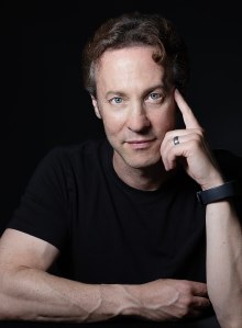 A man with light skin, short curly hair, and blue eyes is wearing a black t-shirt. He is resting his head on his finger, with his arm on a table, and has a calm expression, photographed against a dark background.