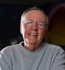 An older man with glasses, short hair, and a light gray sweater smiles while sitting indoors with a dark background.