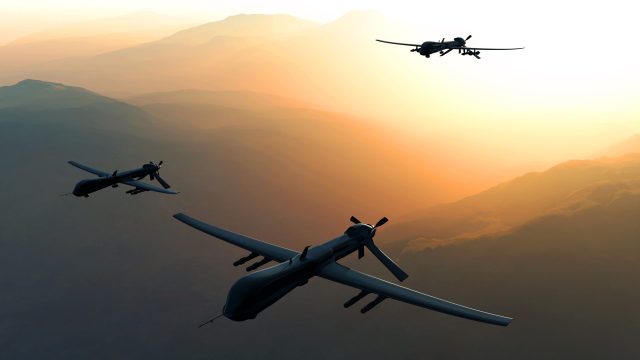 Three drones fly through sky with the sun behind them.