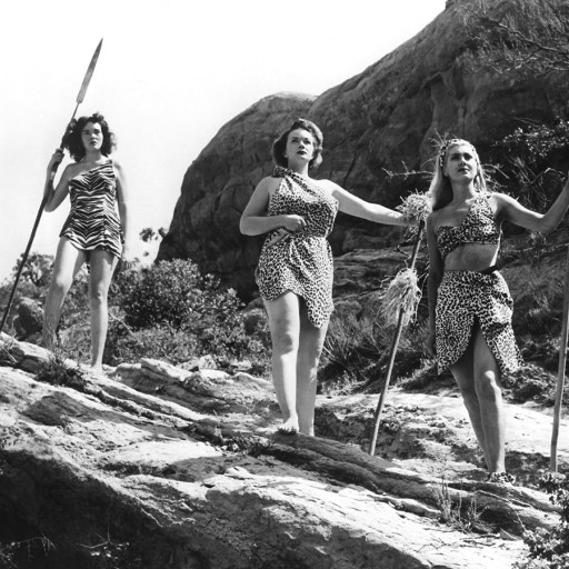 Three women, embodying the male hunter myth, standing confidently on a rock and armed with spears.