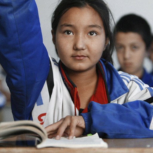 A girl in a blue jacket raises her hand in class, demonstrating language proficiency and recall.