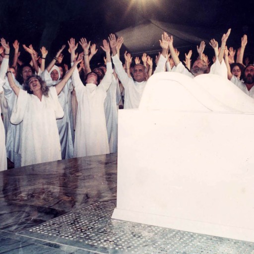 A group of people, part of a doomsday cult, in white robes standing in front of a white chair.
