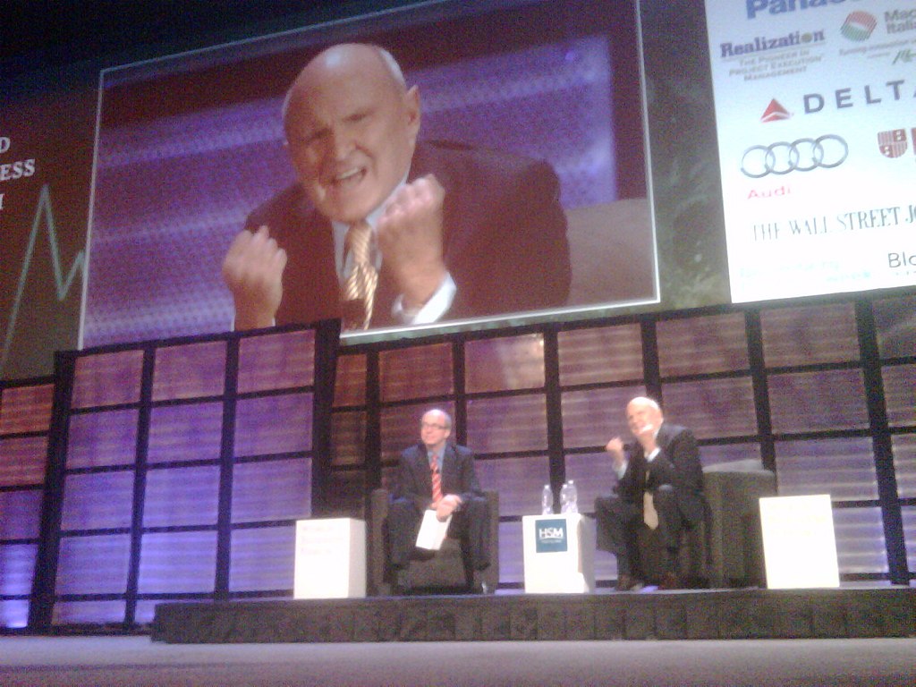 Two men sitting on stage in front of a large screen.