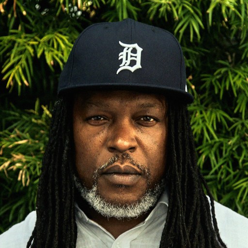 A man with dreadlocks standing in front of a bush.