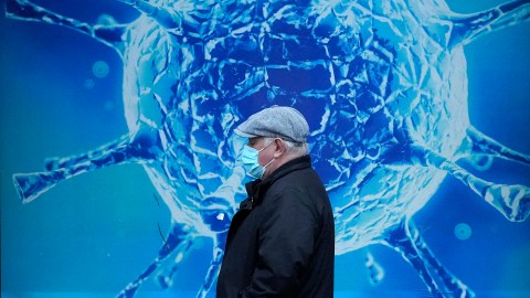 A man wearing a face mask walks past a poster of a coronavirus, symbolizing the ongoing impact of the pandemic.