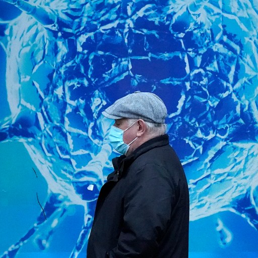 A man wearing a face mask walks past a poster of a coronavirus, symbolizing the ongoing impact of the pandemic.