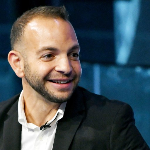 A man in a black suit and white shirt is smiling, unaffected by the doomer mindset.