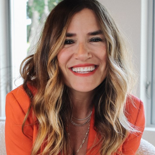A smiling woman in an orange blazer sitting on a couch.