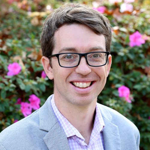 A man in glasses smiles in front of flowers.