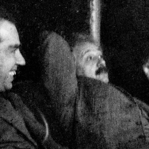 Two men sit closely together, one smiling and the other reclining with a relaxed posture against a dark background.