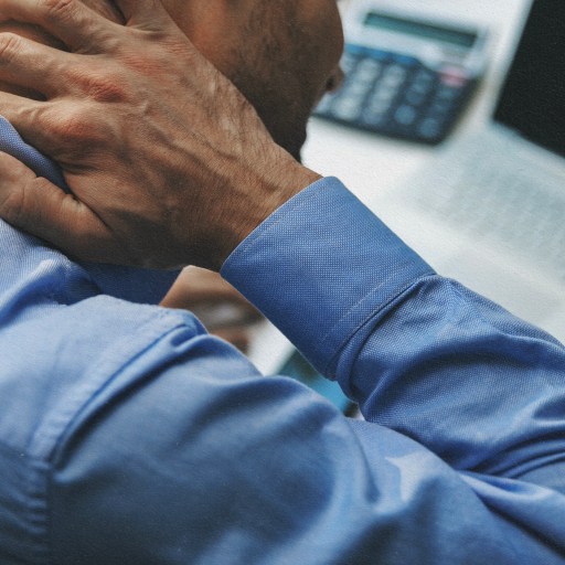 A man in a blue shirt is holding his neck in front of a laptop, possibly experiencing inflammation.