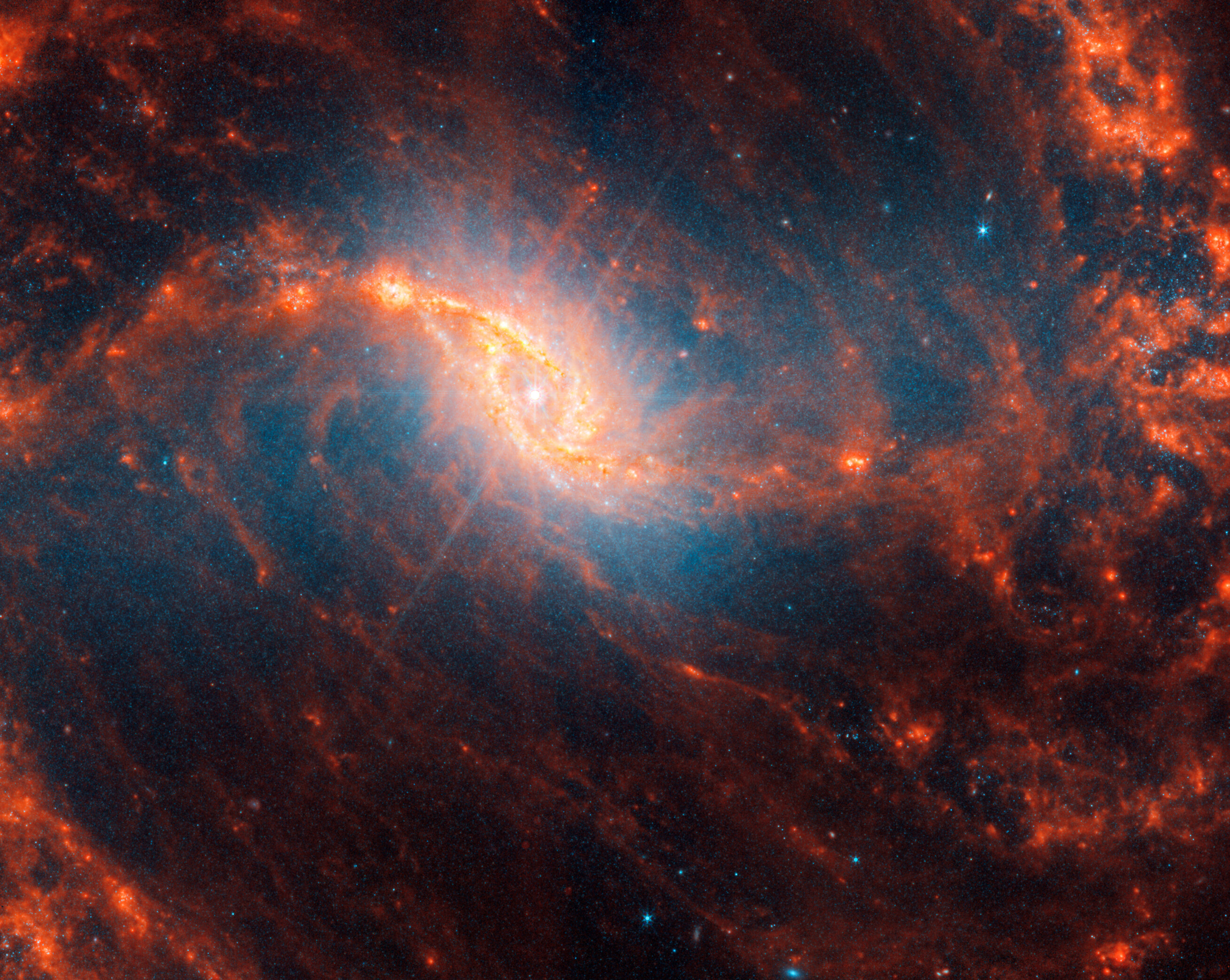 Webb’s image of galaxy NGC 1365 shows a central region that looks like an angled, smashed oval that is a mix of bright orange shades that emanate a blue glow. The galaxy’s bar extends horizontally from the oval. It is crossed by messy dust filaments that curve slightly, forming a backward S shape. Fainter filaments appear below and above the core.