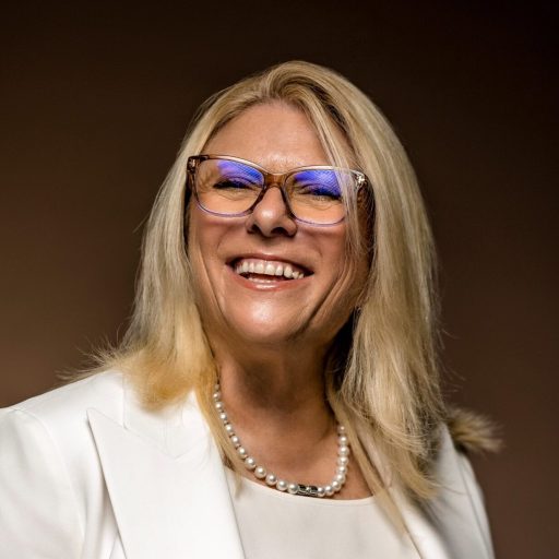 A smiling woman in glasses wearing a white blazer and pearls.