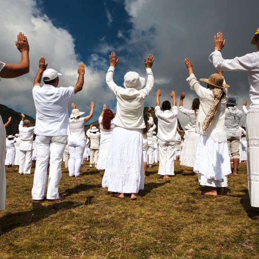 A group of cult members dressed in white participating in an outdoor gathering or ceremony under a partly cloudy sky.