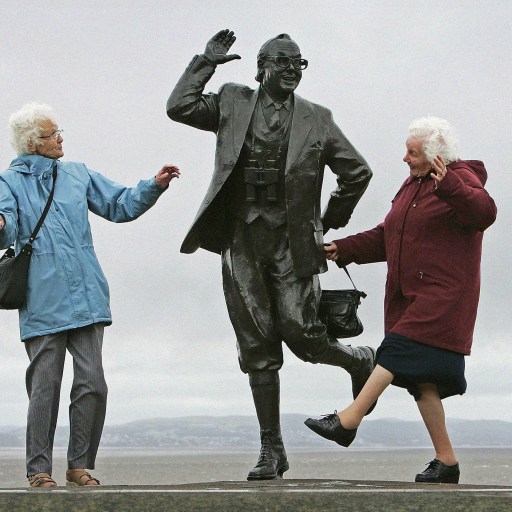 Two elderly women walking alongside and interacting with a statue of a man holding a briefcase.