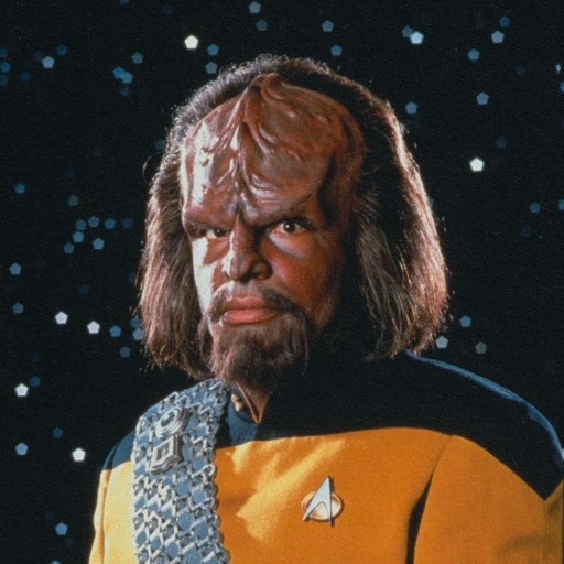 A character in a starfleet uniform with a distinctive ridged forehead, who speaks invented languages, stands against a backdrop of stars.