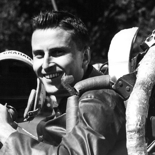 Smiling pilot holding a helmet, wearing a flight suit with a u.s. insignia and a jet pack.