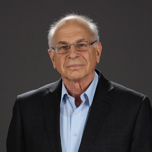 A senior man, Daniel Kahneman, with glasses, wearing a dark suit and a blue shirt, standing against a gray background.