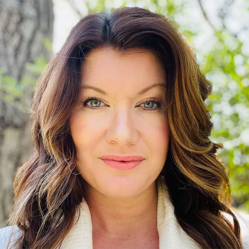 A woman with long, wavy brown hair wearing a white top stands outside in a forested area with green foliage and a tree trunk visible in the background.
