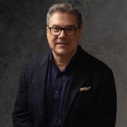 Middle-aged man wearing glasses and a dark blazer, poses against a textured dark gray background.