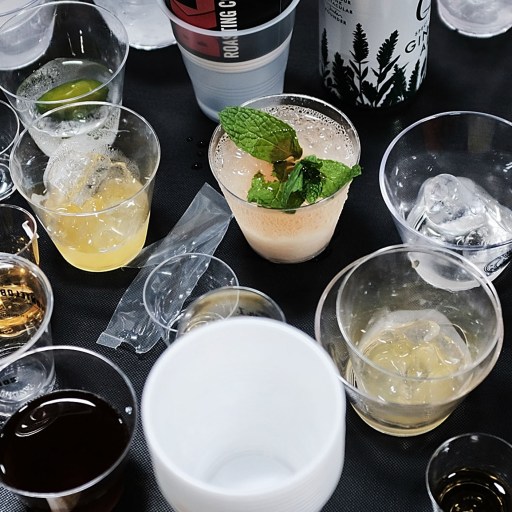 An assortment of half-empty drinks in various plastic cups, some with ice and garnishes, is spread across a dark surface—a telltale sign of the previous night's hangover remedy attempts.