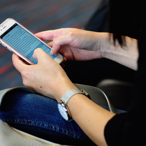 Person seated, using a smartphone to browse social media, with a focus on their hands and the device over a handbag, inside a room with striped carpeting.
