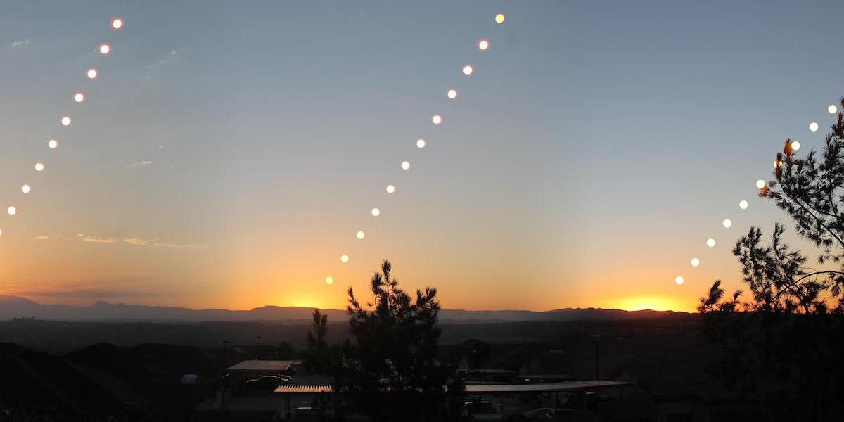 A series of sun positions during sunset over a landscape, with trees in the foreground and mountains in the background, creating a pattern of glowing points in the sky.