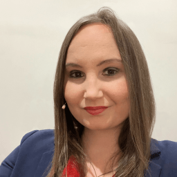 A person with long straight hair, wearing a navy blazer and red shirt, looks at the camera against a plain background. They have a small microphone attached near their left ear.