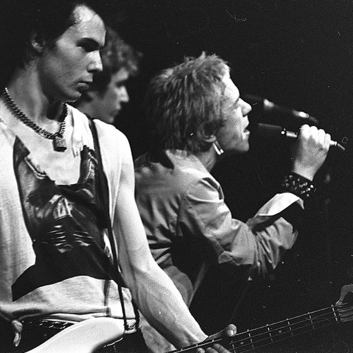 Black and white image of a band performing on stage. One musician is playing a bass guitar while another sings into a microphone. The scene, embodying punk-inspired leaders, has a dark background.