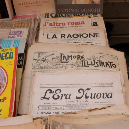 A display of vintage Italian newspapers and magazines, including "Cinema," "La Ragione," and "L'Era Nuova," arranged on a table at an outdoor market.