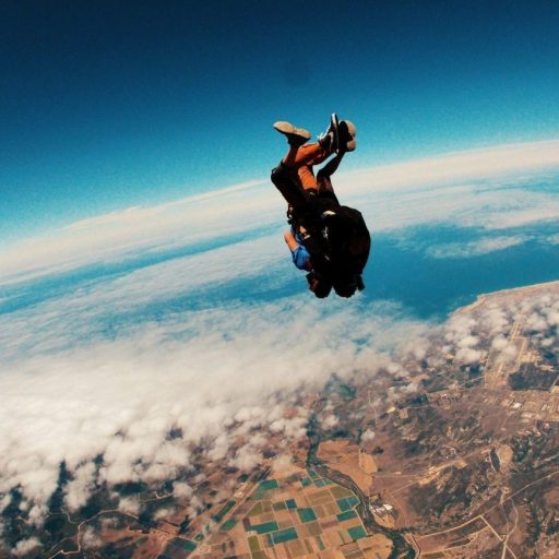 Two people are tandem skydiving, falling through the air above a landscape of fields, mountains, and coastline under a clear sky.