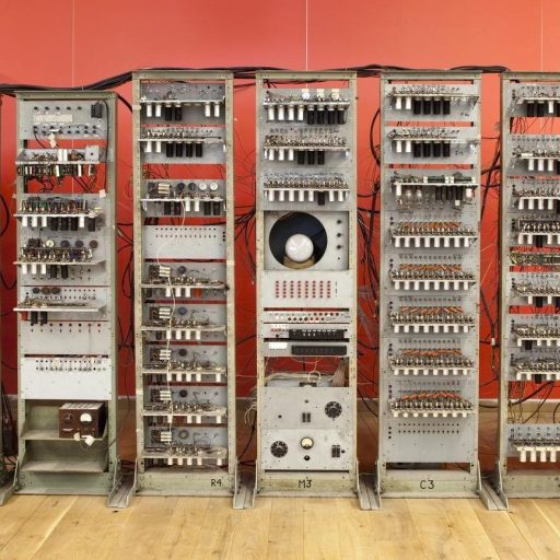 A vintage computer mainframe with multiple interconnected units, cables, and knobs, displayed in front of a red background on a wooden floor.