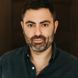 A man with short dark hair and a beard wearing a dark button-up shirt stands against a dark, blurred background, looking directly at the camera.