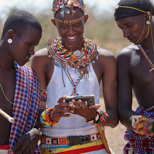Three Masai men, dressed in traditional attire and adorned with beaded jewelry, gather around a smartphone in an outdoor setting.