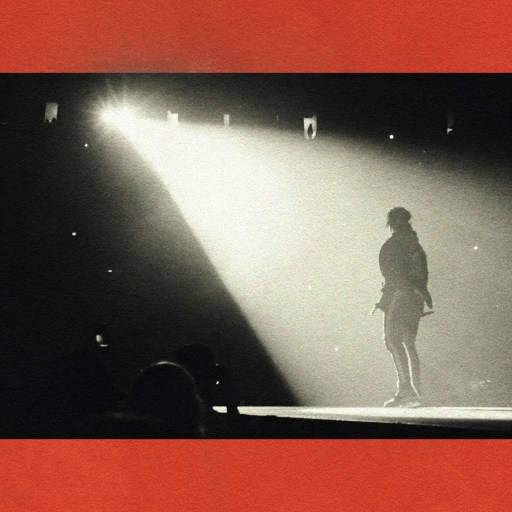Black and white photo of a person on stage, the spotlight effect illuminating them from behind, casting dramatic shadows. A red border frames the image.