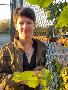 A person with short hair stands outside near a chain-link fence, holding green leaves with the sun casting a soft light.