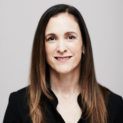 A woman with long, straight brown hair and a black top smiles at the camera against a plain light background.