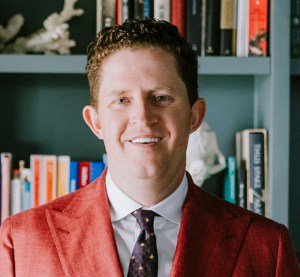 A person with curly hair wearing a red blazer and patterned tie stands in front of a bookshelf filled with books and decorative items.