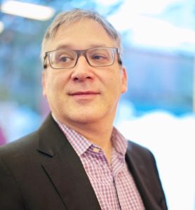 A man with glasses wearing a black blazer and checkered shirt looks to the side with a slight smile.