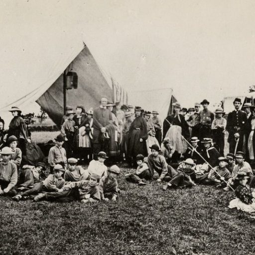 A large group of people, some standing and some sitting, gather outdoors in front of a tent and a horse-drawn carriage, set against a grassy field with a few trees in the background.