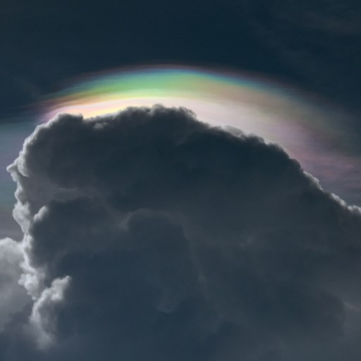 A dark cloud, laced with cynicism, is topped with a colorful iridescent layer against a dim sky.
