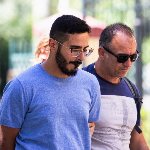 Two men wearing glasses walk side by side outdoors in a sunlit park, with blurred greenery and other people in the background, discussing recent incidents of romance fraud.