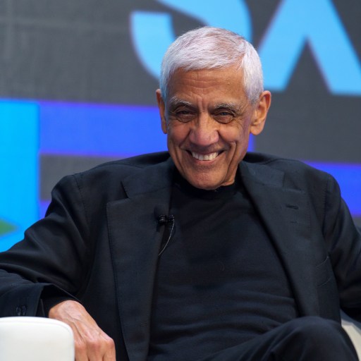 An older man with gray hair is sitting in a white chair, smiling. Dressed in a black outfit, he is framed against a large screen filled with abstract shapes and letters that evoke an AI manifesto.