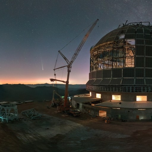 A large telescope observatory under construction at dusk with a visible moon and stars in the sky. Cranes and construction equipment are present around the structure.