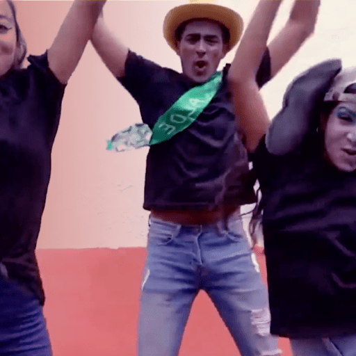 Three people in casual clothing enthusiastically jump in front of a peach-colored wall. One wears a straw hat and a green sash, and another has elephant ears and face paint.