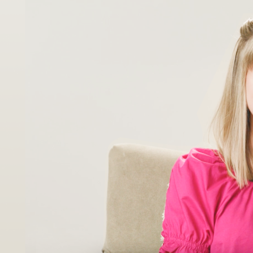 A woman with blonde hair smiles while sitting on a beige chair. She wears a bright pink top and the background is a plain light color.