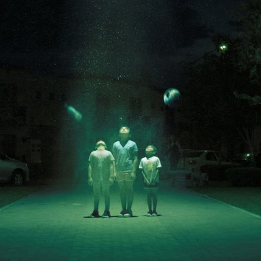Three children stand under a mysterious green light in a suburban street at night, looking upward. Floating objects are illuminated around them.