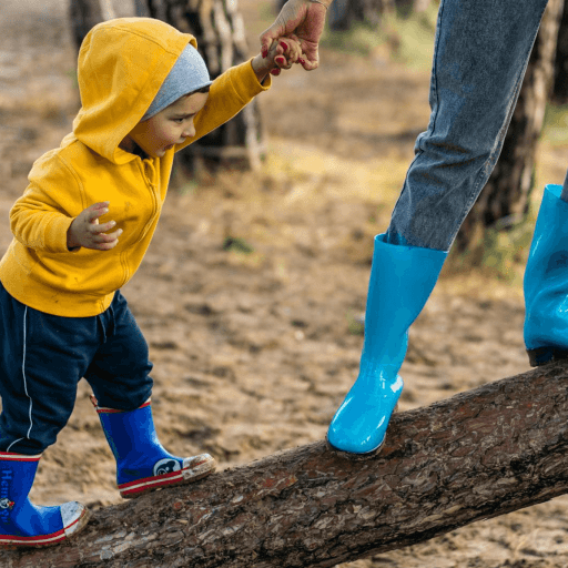 A child in a yellow hoodie and blue boots holds a parent's hand, engaging in play as they walk on a log in a wooded area. The parent, clad in jeans and matching blue boots, guides the little adventurer through nature's playground.